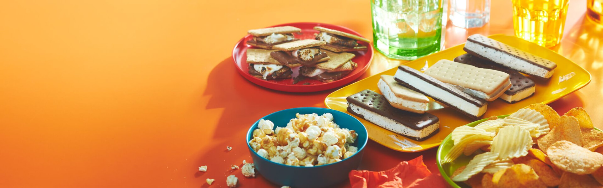 orange background with a plate of ice cream sandwiches, a bowl of chips and popcorn, and a plate of s'mores