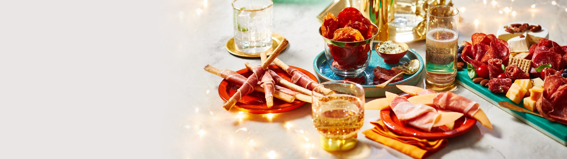 Marble table with twinkle lights, a charcuterie board, plate of salami chips, and prosciutto-wrapped melon and breadsticks.