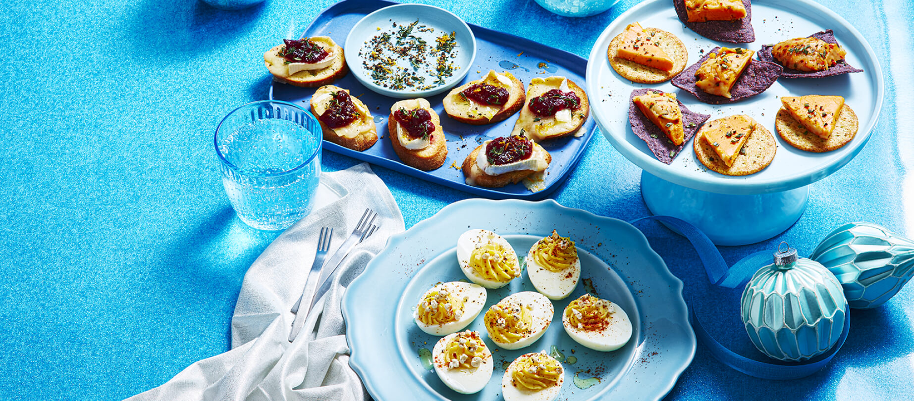 Boule de fromage et croustilles sur un support à gâteau blanc, brie aux canneberges sur un plateau de service bleu et œufs farcis au fromage sur une assiette bleue.