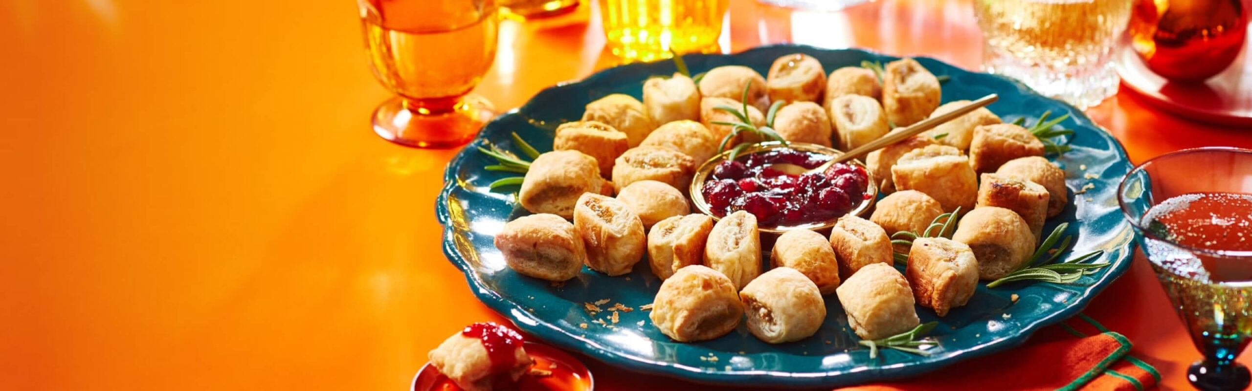 Orange surface with blue plate of sausage rolls arranged in a wreath shape with fresh rosemary and dipping sauce