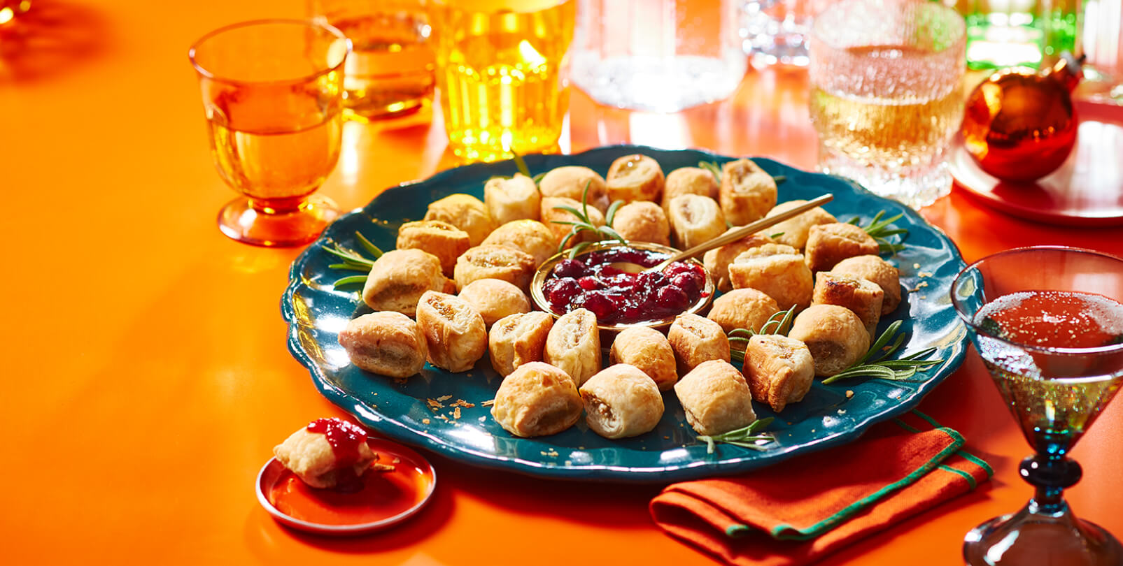 Une assiette bleue de rouleaux de saucisses disposés en forme de couronne avec du romarin frais et une sauce à tremper.