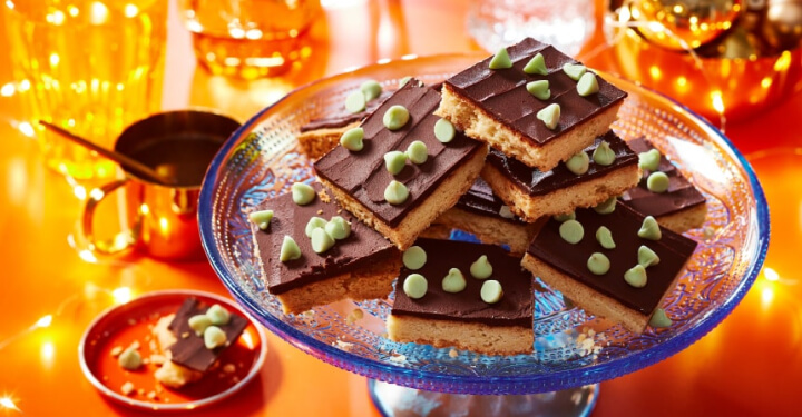 Orange table with twinkle light décor, and a blue glass cake stand topped with mint chocolate shortbread slice cookies with mint flavoured baking chips sprinkled over the top