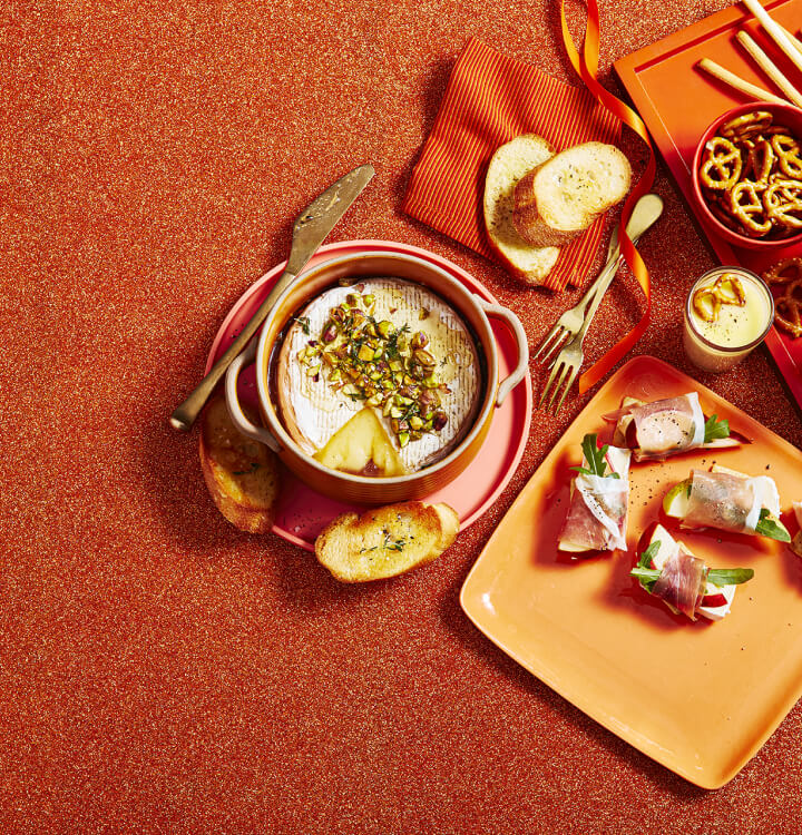 Baked brie topped with crushed nuts and a side of crostinis on a pink plate, and brie and pear bites on an orange serving tray.